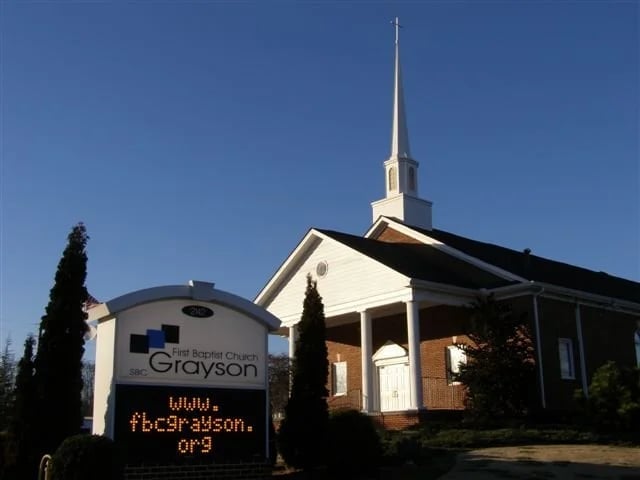 Front view of a brick church building with white steeple and a sign reading First Baptist Church Grayson against a clear blue sky